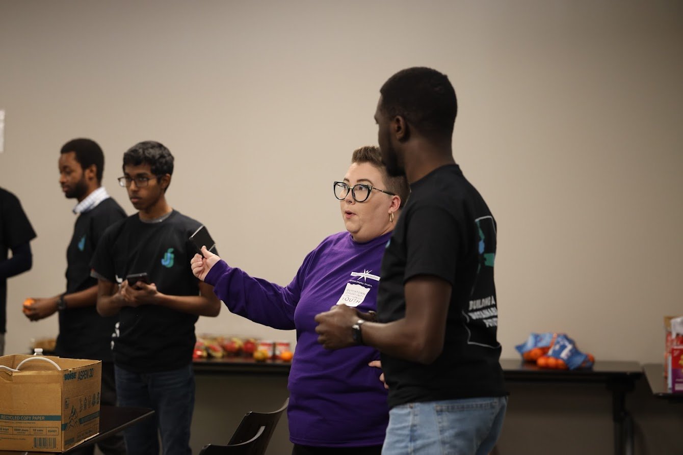 Two people in conversation at an indoor community event, with team members in JustUsedTech shirts and supplies laid out on tables behind them. (20+ active volunteers)