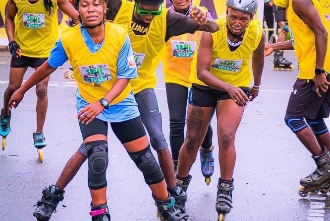 Four skaters in yellow event bibs, helmets, and knee pads skating together along a road.