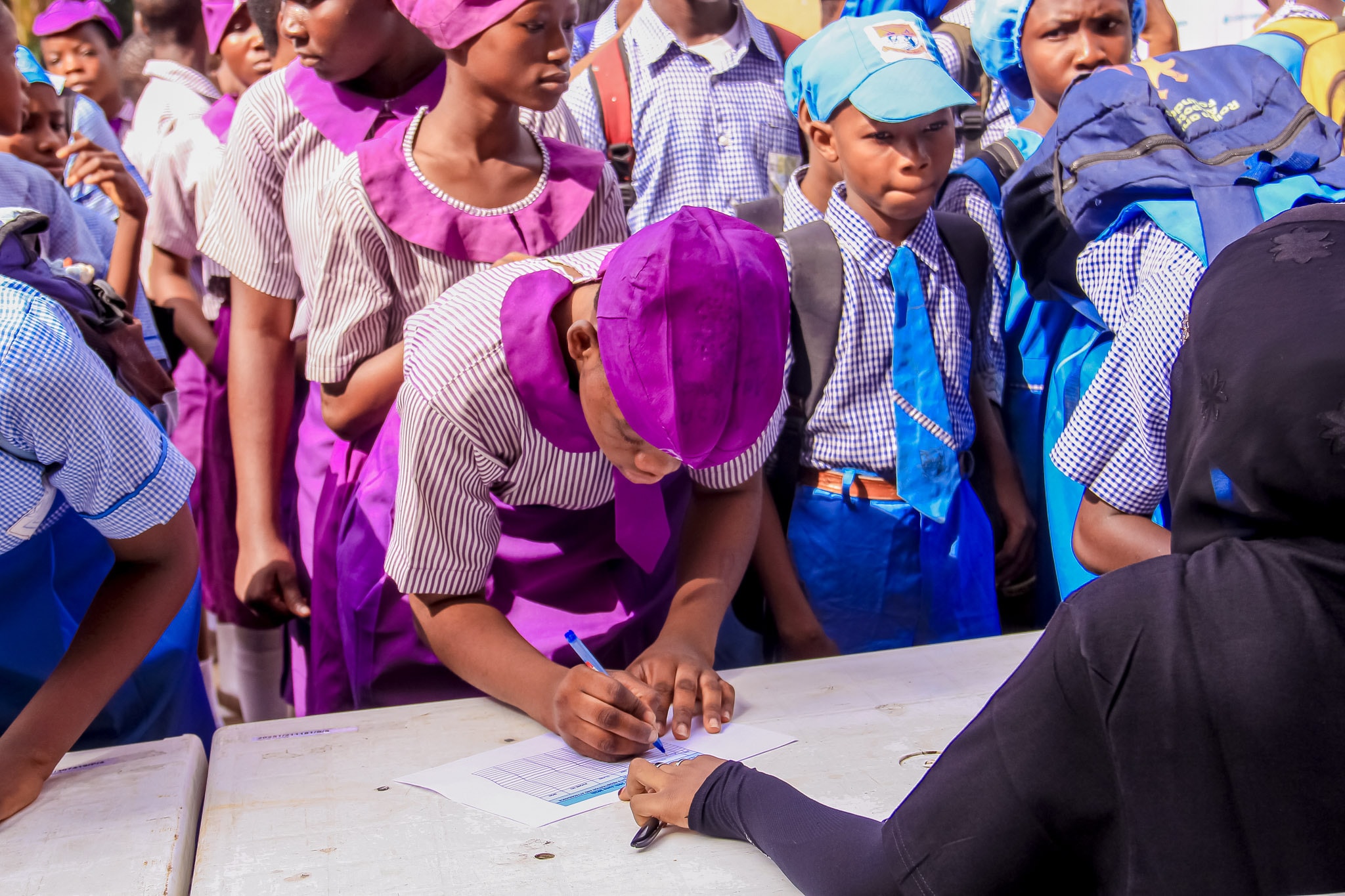 Students in school uniform crowded around a registration table while a volunteer writes on a form.