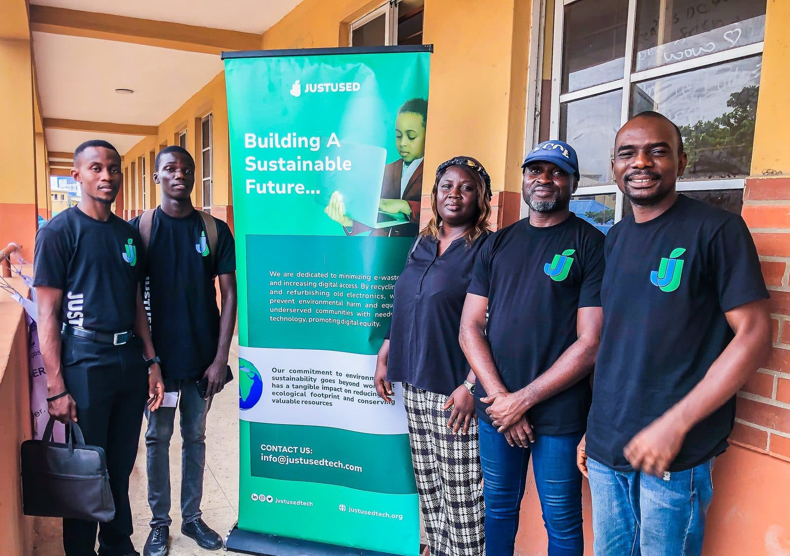 Five team members in JustUsedTech shirts standing beside a Building A Sustainable Future banner on a school walkway.