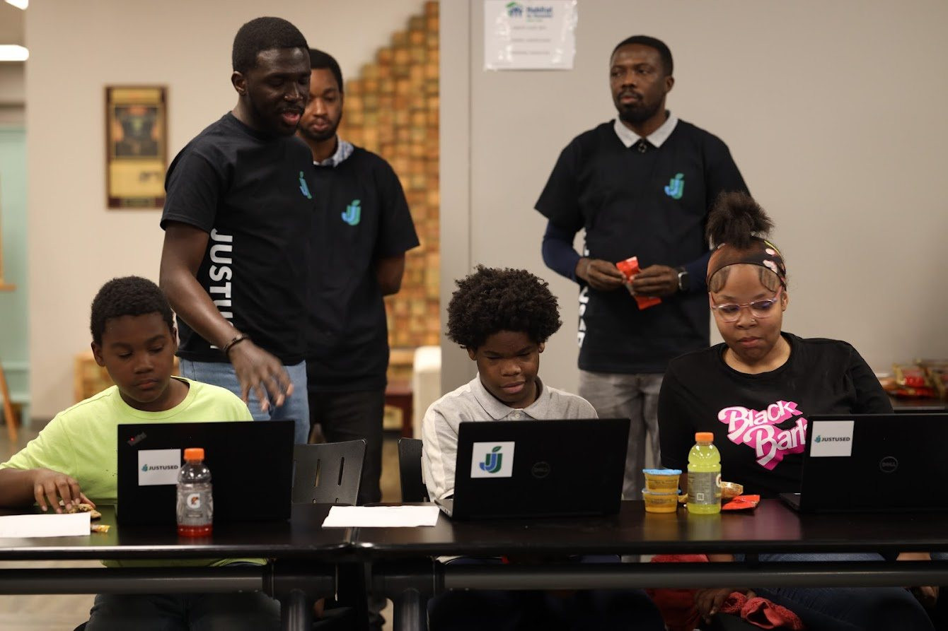 Young people working at laptops around a table, with team members in JustUsedTech shirts standing alongside.