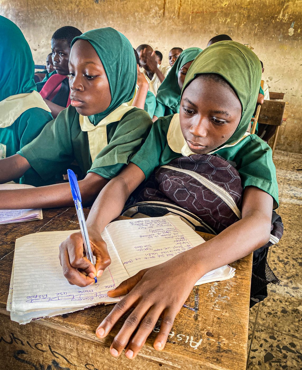 Two students in school uniform writing in an exercise book at a classroom desk.